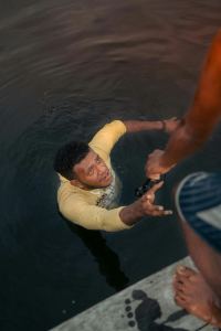 A man in a yellow shirt is in the water reaching up, while another person on a dock reaches down to help him. - PsychDiscouse