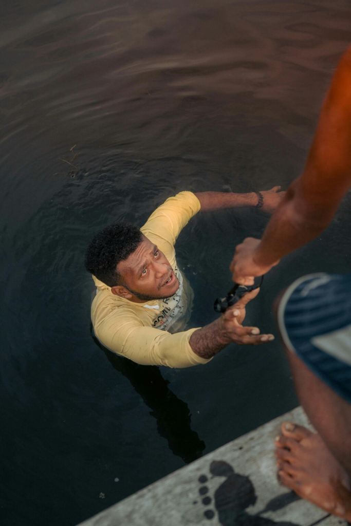 A man in a yellow shirt is in the water reaching up, while another person on a dock reaches down to help him. - PsychDiscouse