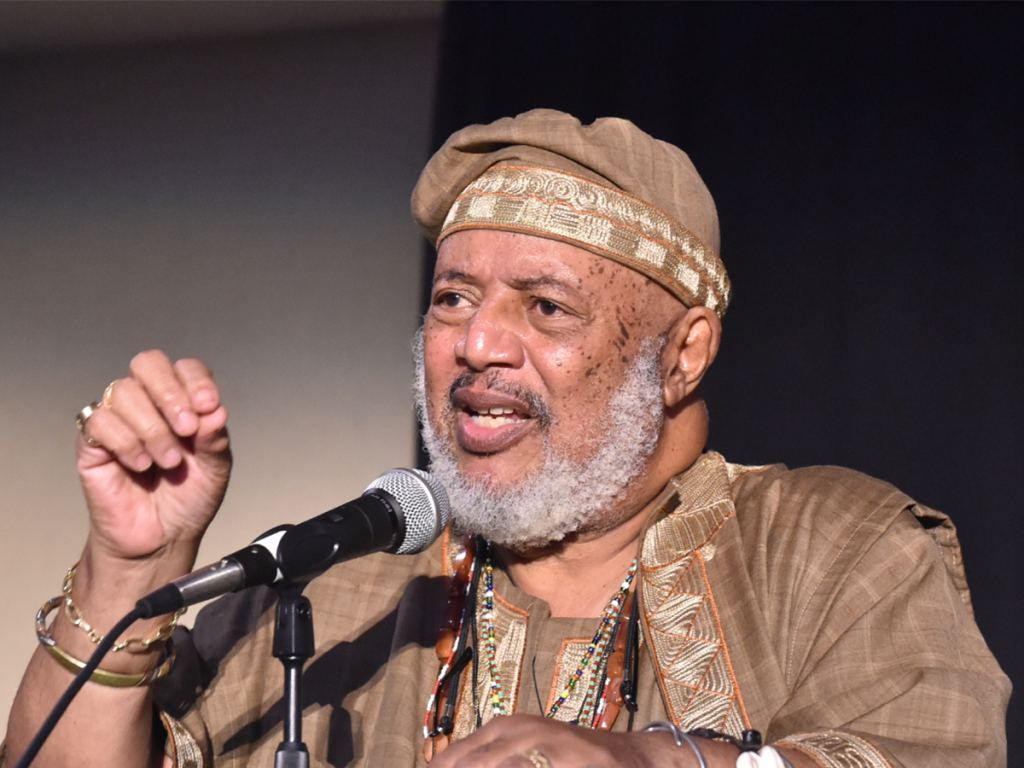 An older man with a gray beard, wearing traditional brown clothing and a matching hat, speaks into a microphone while gesturing with his hand. - PsychDiscouse