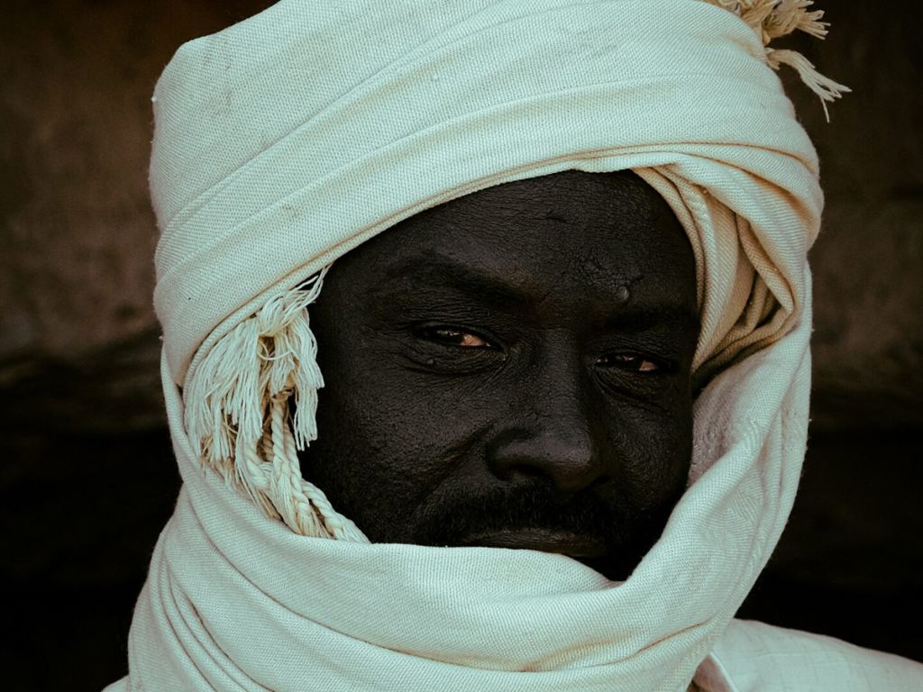A man with dark skin wears a white turban and scarf, with only his face partially visible, looking directly at the camera. - PsychDiscouse