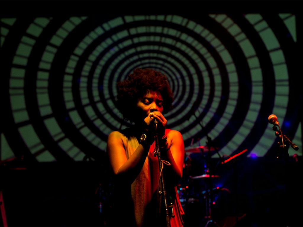 A singer performs on stage in front of a large, circular, hypnotic pattern projected in the background, with microphone stands and dim stage lighting. - PsychDiscouse