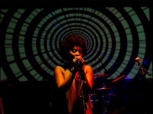 A singer performs on stage in front of a large, circular, hypnotic pattern projected in the background, with microphone stands and dim stage lighting. - PsychDiscouse