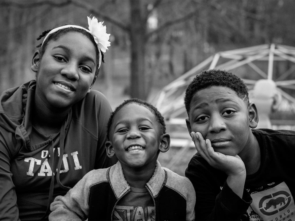 Three children sit close together outdoors, smiling and looking at the camera; the background is blurred with trees and a dome structure visible. - PsychDiscouse