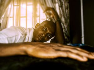 A person lies on a bed near a sunlit window, looking directly at the camera with a neutral expression, one arm extended toward the foreground. - PsychDiscouse