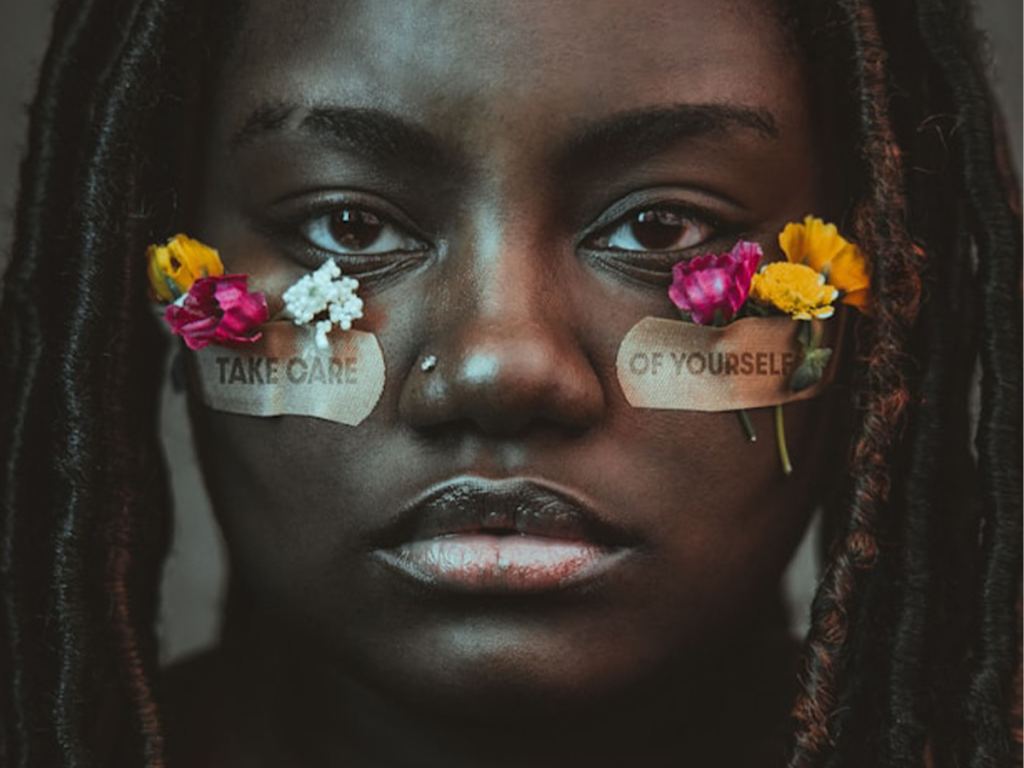 A person with flowers tucked near their eyes wears two bandages under each eye reading "TAKE CARE" and "OF YOURSELF. - PsychDiscouse
