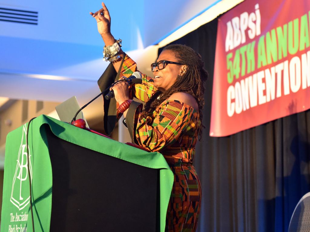 A woman in a patterned dress speaks at a podium, raising her right arm, during the ABPsi 54th Annual Convention. - PsychDiscouse