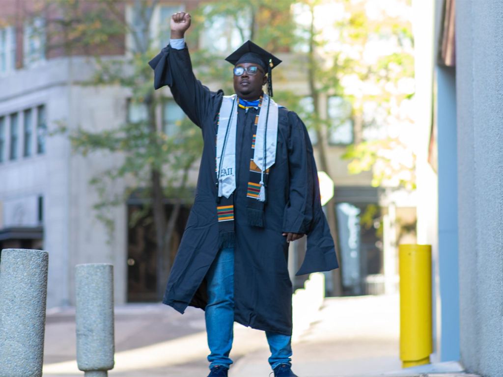 A graduate wearing a cap, gown, and sash stands outdoors with one fist raised in the air. - PsychDiscouse