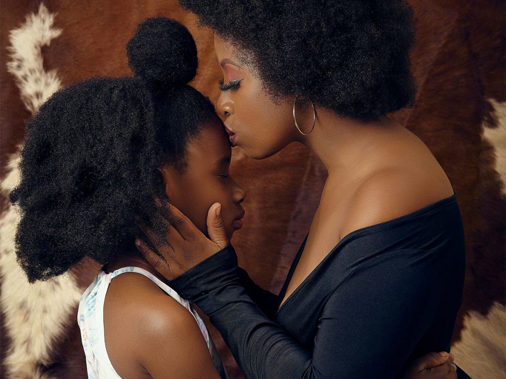 An adult embraces and kisses the forehead of a child, both with closed eyes, in front of a brown and white patterned background. - PsychDiscouse