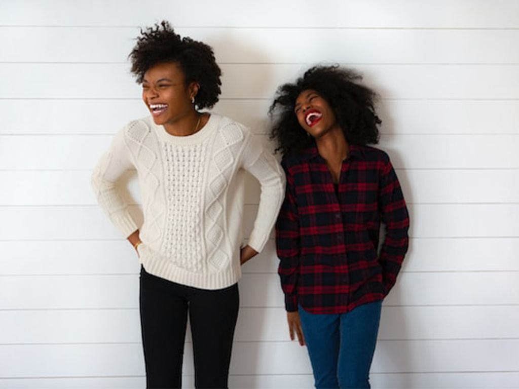 Two women standing against a white shiplap wall, smiling and laughing. One wears a white sweater and black pants, the other a red plaid shirt and blue jeans. - PsychDiscouse