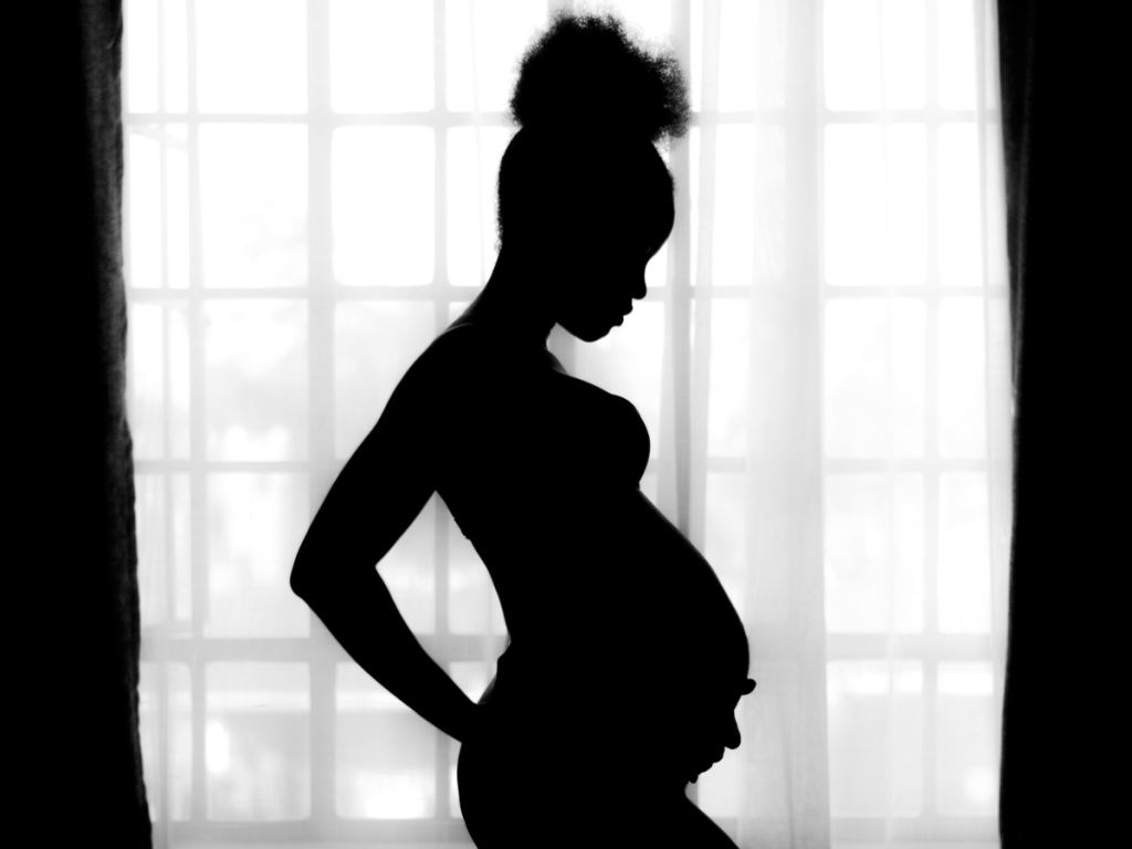 Silhouette of a pregnant woman standing in profile by a window with bright light streaming through sheer curtains. - PsychDiscouse