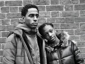 Two people in winter jackets stand close together against a brick wall, with one resting their head on the other's shoulder. Both have neutral expressions. Black and white photo. - PsychDiscouse
