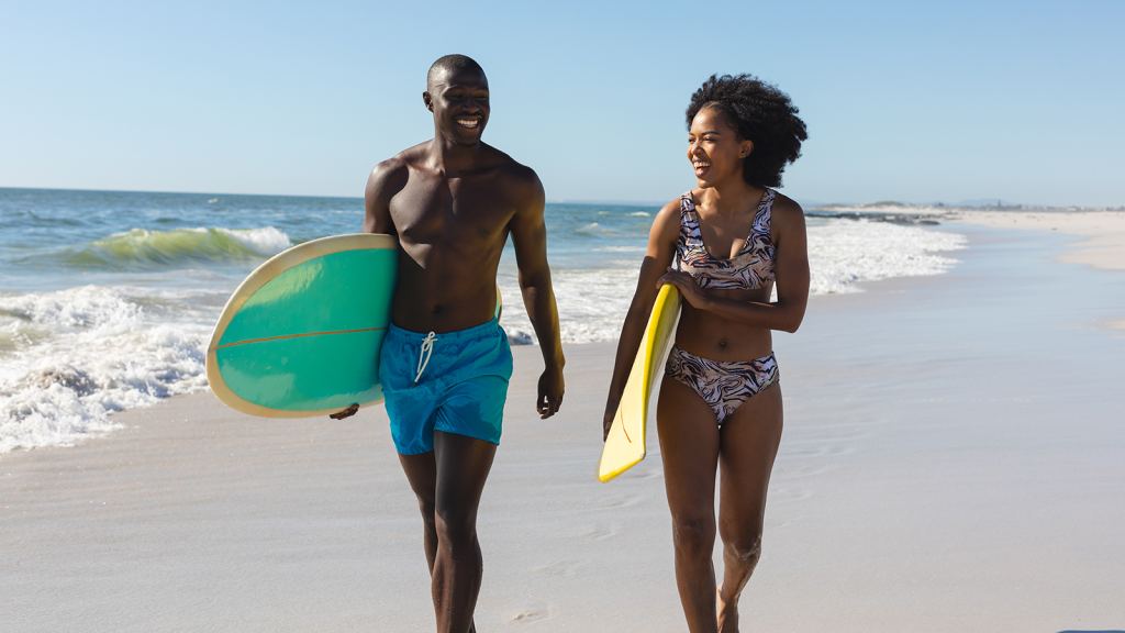 A man and woman in swimwear walk along the beach carrying surfboards, smiling and talking with the ocean in the background. - PsychDiscouse