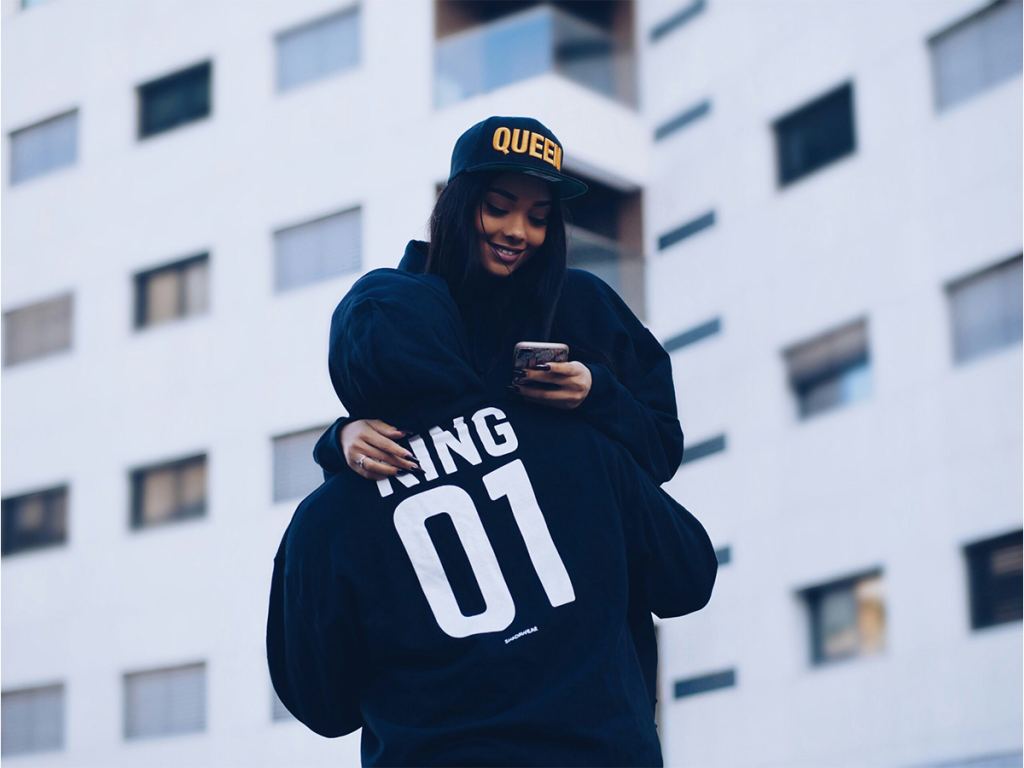 A person wearing a "KING 01" hoodie hugs a smiling person in a "QUEEN" hat, who is looking at a smartphone, with a modern building in the background. - PsychDiscouse