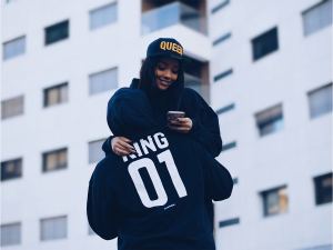 A person wearing a "KING 01" hoodie hugs a smiling person in a "QUEEN" hat, who is looking at a smartphone, with a modern building in the background. - PsychDiscouse