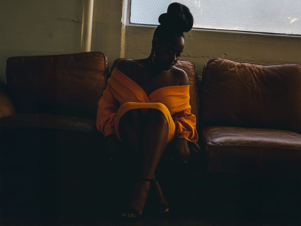 A woman in an off-shoulder yellow dress sits on a brown leather couch in a dimly lit room near a window. - PsychDiscouse