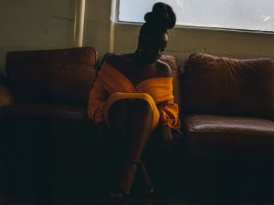 A woman in an off-shoulder yellow dress sits on a brown leather couch in a dimly lit room near a window. - PsychDiscouse