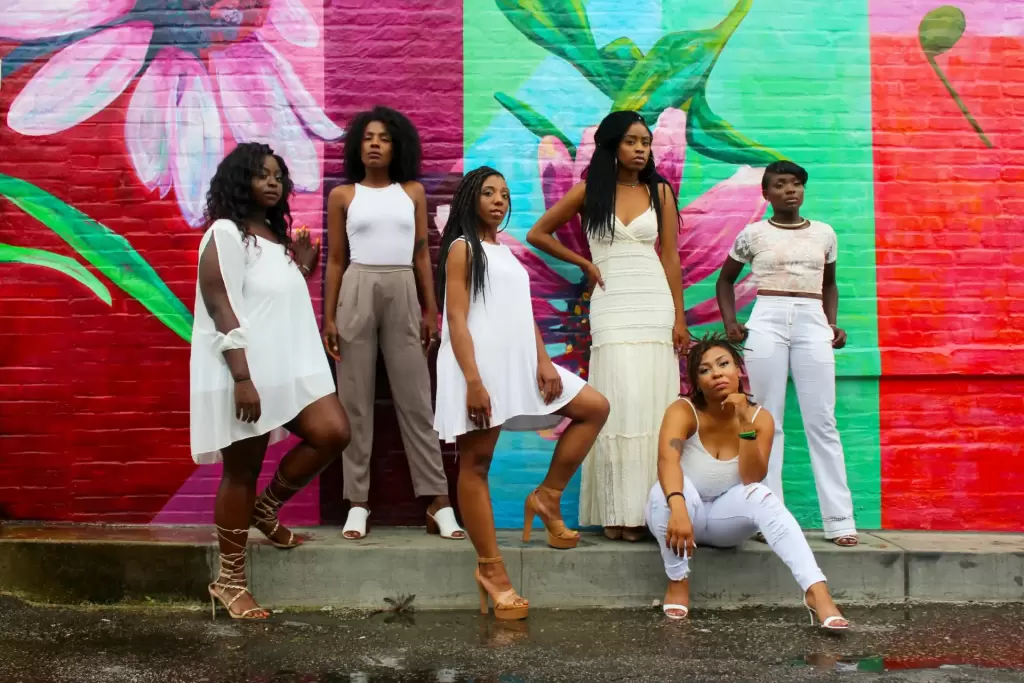 Six women pose confidently in front of a colorful floral mural, wearing stylish white and neutral-toned outfits and heels. - PsychDiscouse