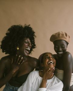 Three women smiling and laughing together in front of a beige background, wearing stylish outfits and accessories. - PsychDiscouse