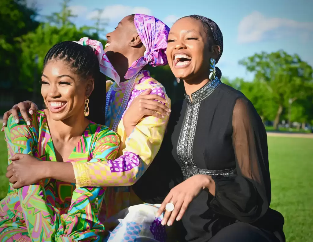 Three women sit on grass outdoors, smiling and laughing together in colorful, patterned outfits on a sunny day, with trees and blue sky in the background. - PsychDiscouse
