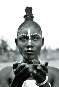 A person with white face paint and jewelry holds a smoking bowl, gazing forward. The background is blurred, and the image is in black and white. - PsychDiscouse
