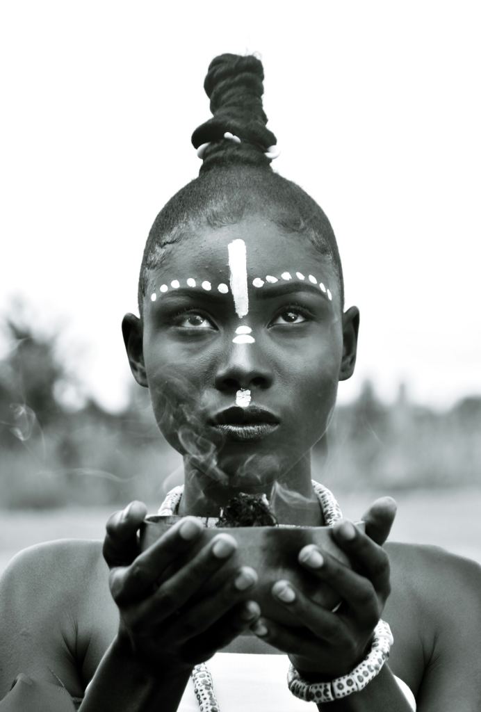 A person with white face paint and jewelry holds a smoking bowl, gazing forward. The background is blurred, and the image is in black and white. - PsychDiscouse