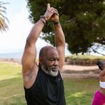 Two people practice yoga outdoors on grass, each stretching with arms raised overhead. Trees and a palm tree are visible in the background. - PsychDiscouse