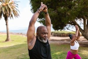 Two people practice yoga outdoors on grass, each stretching with arms raised overhead. Trees and a palm tree are visible in the background. - PsychDiscouse