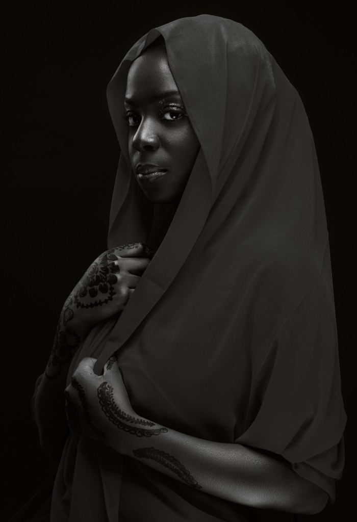 A woman with henna-decorated hands wears a draped, dark shawl over her head and shoulders, gazing forward with a calm expression against a black background. - PsychDiscouse