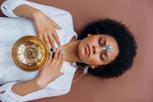 A person lies on their back with closed eyes, holding a brass singing bowl on their chest and sensors attached to their forehead, suggesting a meditation or relaxation session. - PsychDiscouse