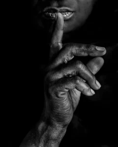 Black and white close-up of a person holding a finger to their lips in a gesture for silence. - PsychDiscouse