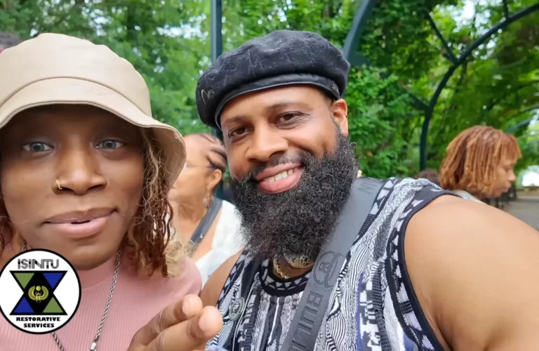 A woman in a bucket hat and a man with a beard and black beret pose for a selfie in a park, with an “ISINTU Restorative Services” logo in the corner. - PsychDiscouse