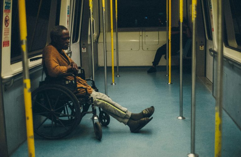 An older person in a wheelchair sits alone on a mostly empty subway car with yellow poles and blue floors. Another person sits in the background. - PsychDiscouse