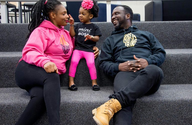 A woman, a man, and a young girl sit together on carpeted stairs, smiling and interacting. The child wears a “mini boss” shirt and a pink flower headband. - PsychDiscouse