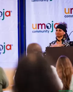 A woman stands at a podium speaking at an event, with an “Umoja Community” banner in the background and audience members visible in the foreground. - PsychDiscouse