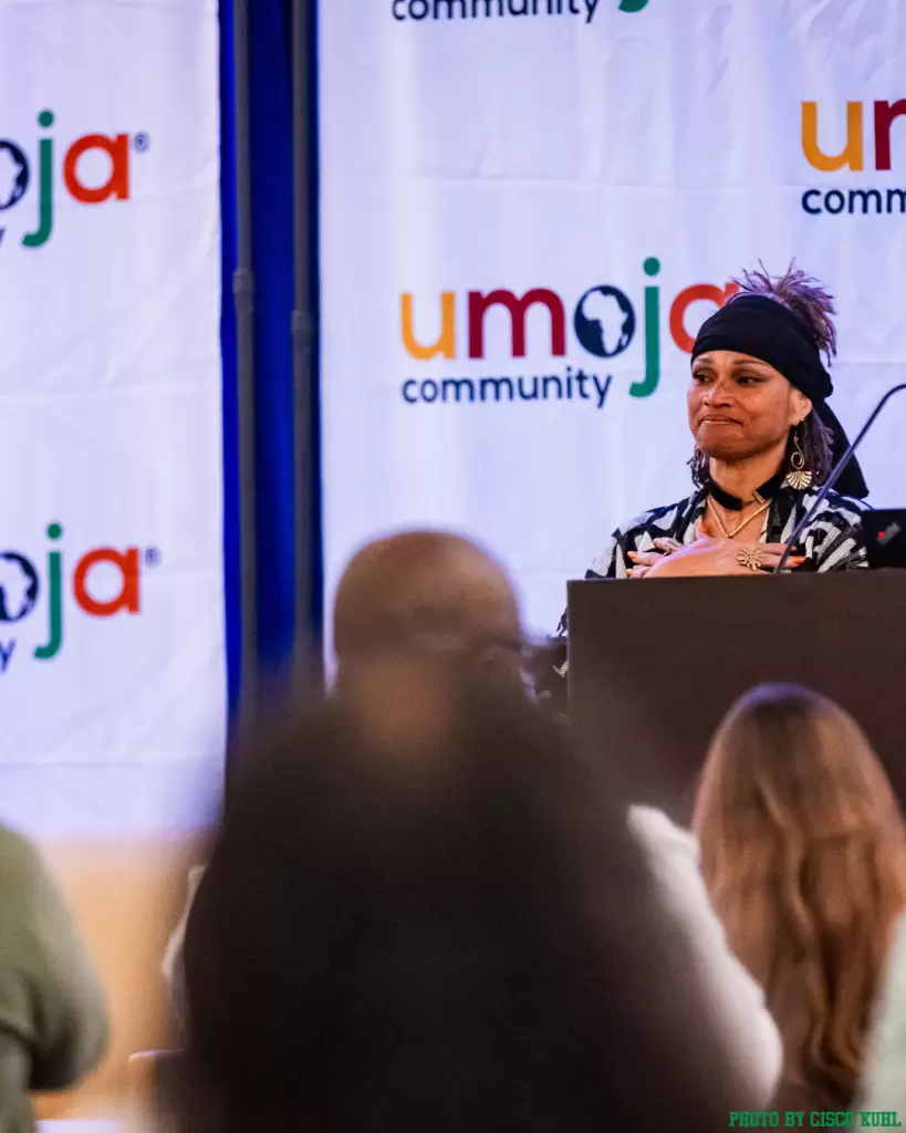 A woman stands at a podium speaking at an event, with an “Umoja Community” banner in the background and audience members visible in the foreground. - PsychDiscouse