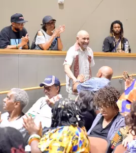 A man stands and smiles as people around him applaud in a crowded auditorium; some in balcony seats also clap and watch the scene below. - PsychDiscouse