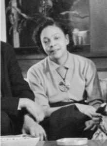 A woman sits indoors, wearing a collared shirt and pendant necklace, with her hands resting on her lap. Part of another person is visible next to her. - PsychDiscouse