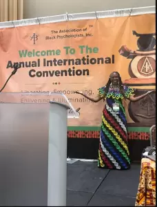 A woman in a colorful dress stands on stage, smiling, in front of a banner for The Association of Black Psychologists’ Annual International Convention. - PsychDiscouse
