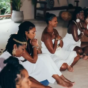 A group of women dressed in white sit on the floor with eyes closed, hands clasped in front of them, participating in a meditative or spiritual practice indoors. - PsychDiscouse