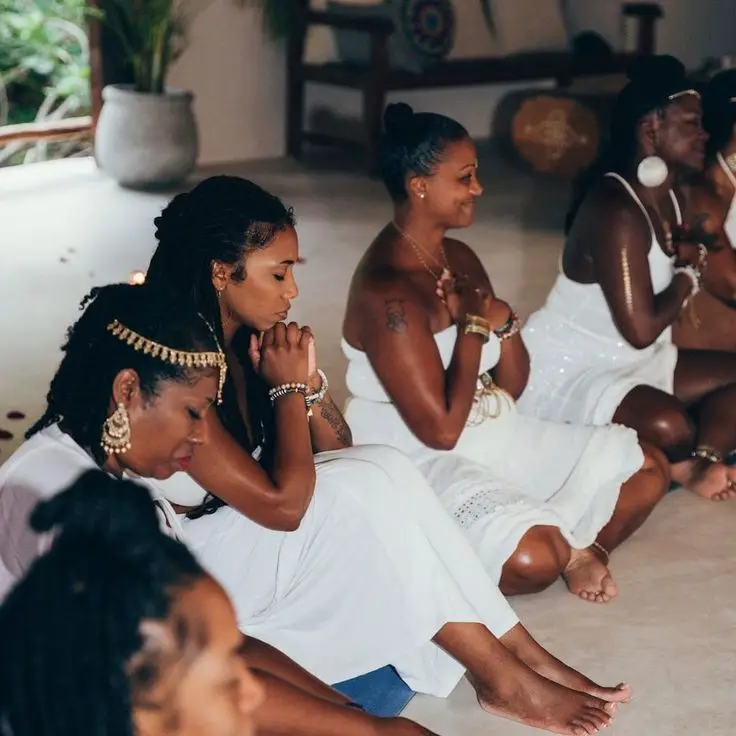 A group of women dressed in white sit on the floor with eyes closed, hands clasped in front of them, participating in a meditative or spiritual practice indoors. - PsychDiscouse