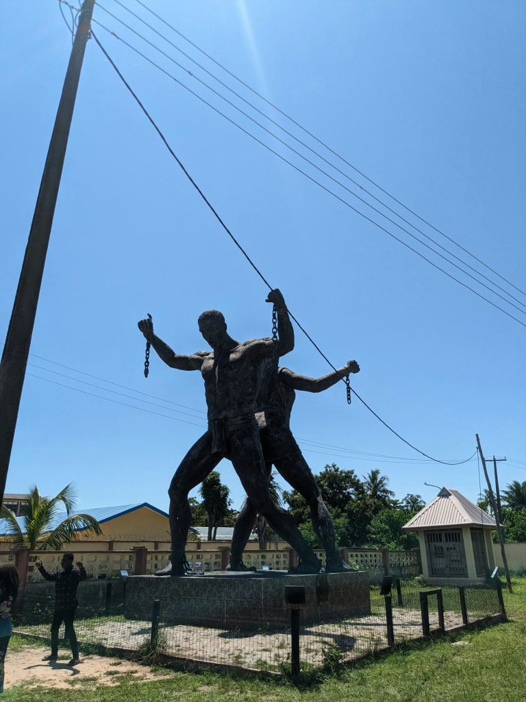 Bronze sculpture of two figures holding broken chains, standing on a raised platform outdoors, with buildings, trees, and a clear blue sky in the background. - PsychDiscouse