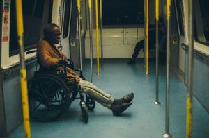 An older person in a wheelchair sits alone on a mostly empty subway car with yellow poles and blue floors. Another person sits in the background. - PsychDiscouse