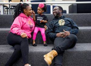 A woman, a man, and a young girl sit together on carpeted stairs, smiling and interacting. The child wears a “mini boss” shirt and a pink flower headband. - PsychDiscouse