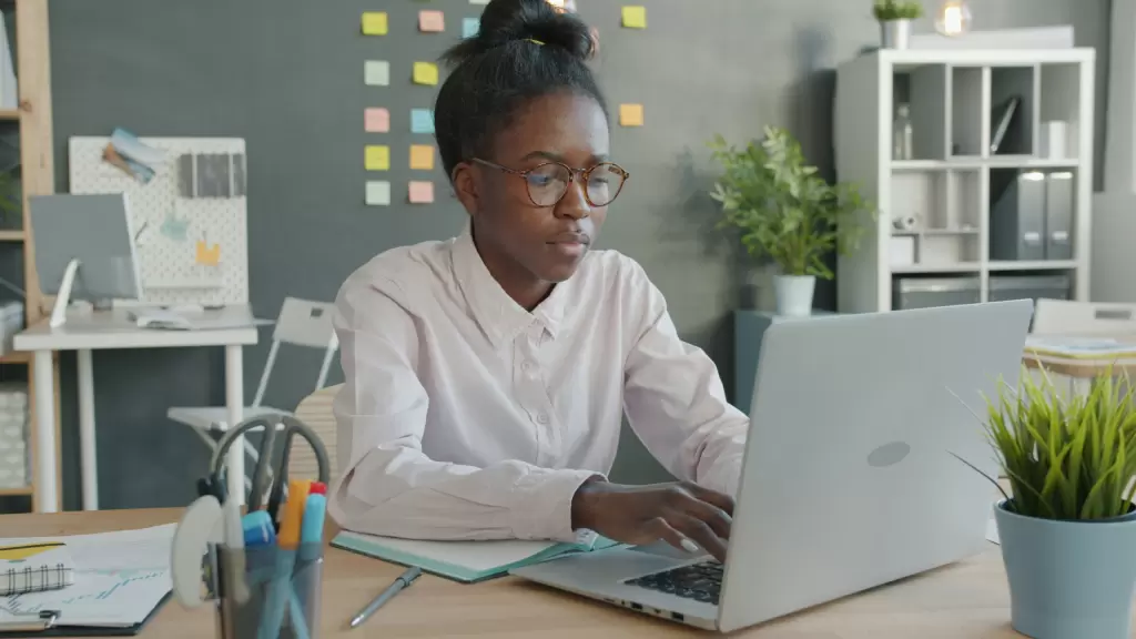 Person wearing glasses works on a laptop at a desk in a modern office with sticky notes on the wall and office supplies nearby. - PsychDiscouse