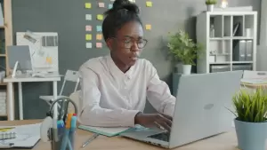Person wearing glasses works on a laptop at a desk in a modern office with sticky notes on the wall and office supplies nearby. - PsychDiscouse