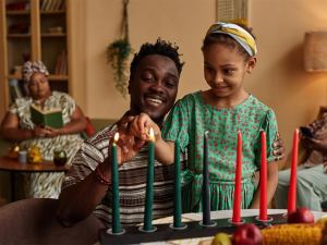 A man and a young girl light a green candle on a kinara for Kwanzaa, with two women sitting in the background. - PsychDiscouse