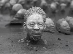 A realistic sculpture of a man's head and shoulders partially buried in soil, with similar heads in the background; the image is in black and white. - PsychDiscouse