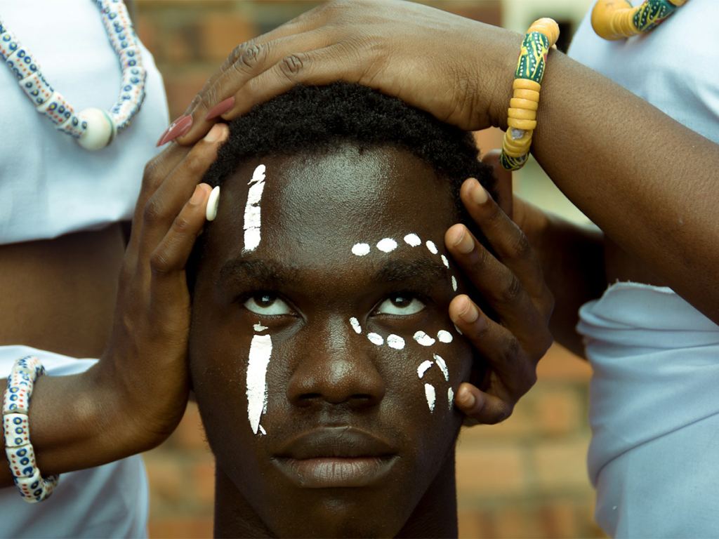 A young person with white face paint looks upward as hands adorned with beaded jewelry rest gently on their head against a blurred background. - PsychDiscouse