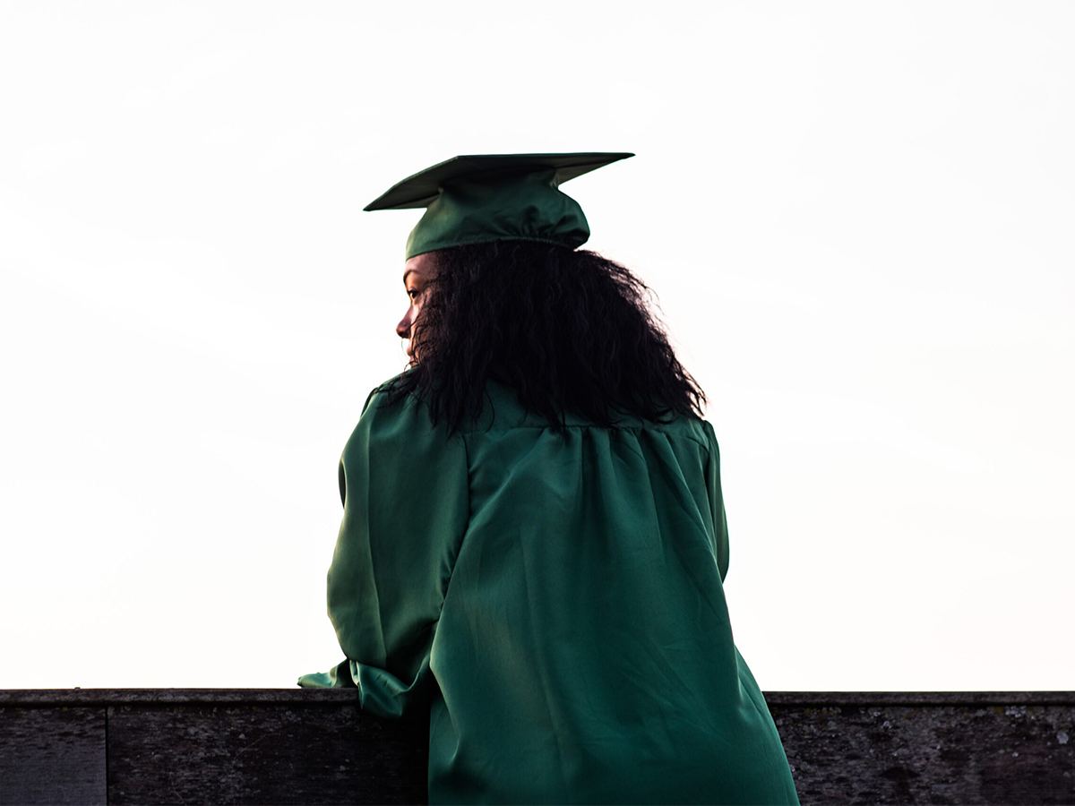 Person in a green graduation cap and gown stands outdoors, facing away from the camera, looking into the distance. - PsychDiscouse