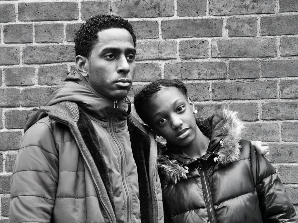 Two people in winter jackets stand close together against a brick wall, with one resting their head on the other's shoulder. Both have neutral expressions. Black and white photo. - PsychDiscouse